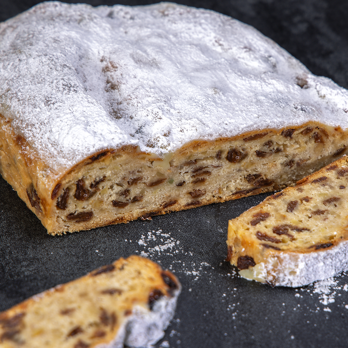 Traditioneller Chris-Stollen als Rosinenstollen mit Puderzucker und saftigen Rosinen, angeschnitten auf dunklem Untergrund.